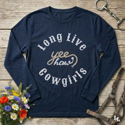 A navy blue long-sleeve graphic t-shirt laid flat on a rustic wooden surface, surrounded by cowboy-themed props including a halter, horse bit, and wildflowers. The shirt features a graphic print reading 'Long Live yeehaw Cowgirls' in white and glittery gold lettering.