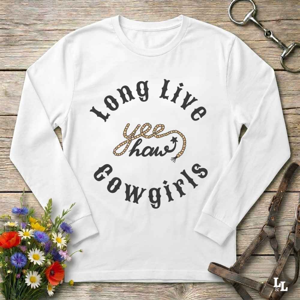 A white long-sleeve t-shirt with 'Long Live yee haw cowgirls' printed on the front, styled in a flat-lay on a rustic wooden background with equestrian and floral props.