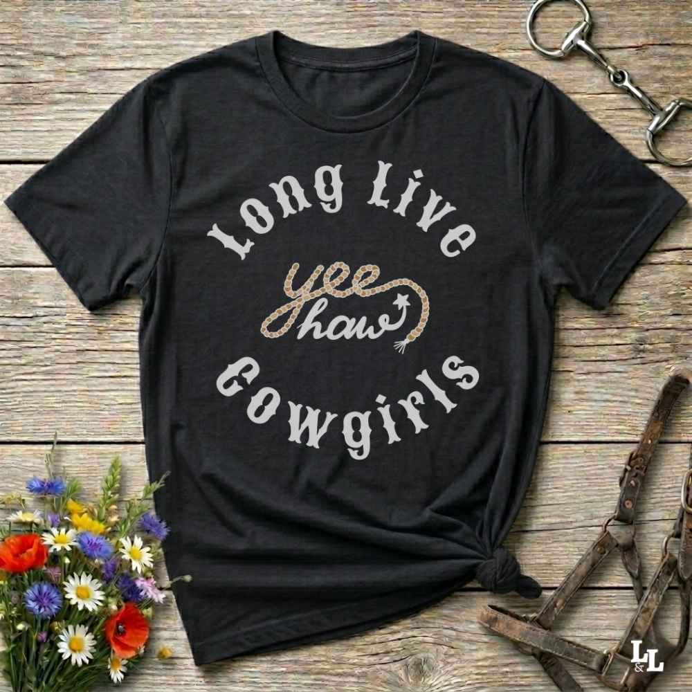 A black graphic t-shirt with the text 'Long Live yee haw cowgirls' in white and glittery gold, styled in a flat-lay on a rustic wooden background with equestrian accessories and flowers.