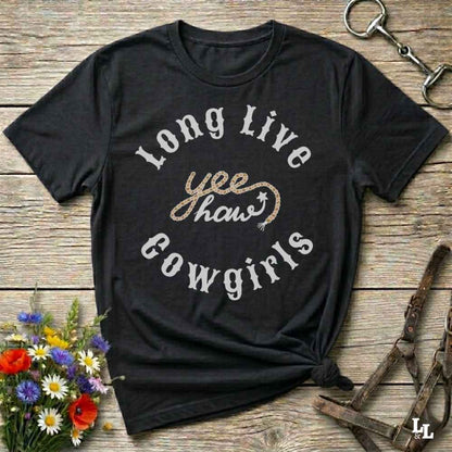 A black graphic t-shirt with the text 'Long Live yee haw cowgirls' in white and glittery gold, styled in a flat-lay on a rustic wooden background with equestrian accessories and flowers.