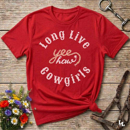 A red short-sleeve graphic T-shirt laid flat on a rustic wooden surface, with white and glittery gold text reading 'Long Live yee haws cowgirls.' Decorative flowers and horse tack are placed around it as styling props.