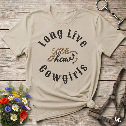A beige graphic t-shirt with 'Long Live yee haws cowgirls' printed on the front, styled in a flat-lay on a rustic wooden background with a horse bit, halter, and wildflowers.