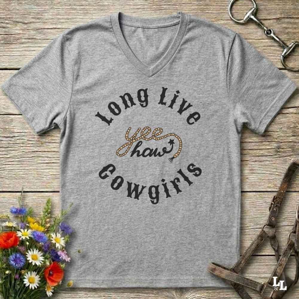 A heather gray V-neck t-shirt with a western-themed graphic reading 'Long Live yee haw! Cowgirls' in black and gold, displayed on a rustic wooden background with flowers and horse tack props.