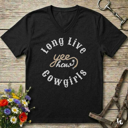 A black V-neck t-shirt with a western-themed graphic reading 'Long Live yee haws Cowgirls' in white and gold, displayed on a rustic wooden background with equestrian accessories and flowers.