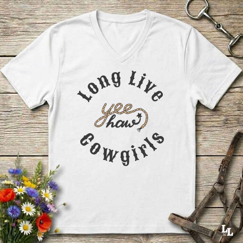 A white V-neck t-shirt with a western-themed graphic reading 'Long Live yee haws Cowgirls' in black and leopard-print lettering, displayed flat on a rustic wooden background with equestrian and floral props.