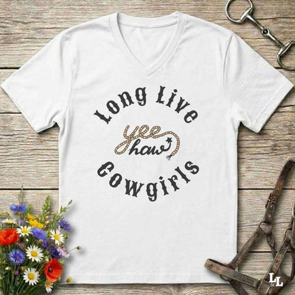 A white V-neck t-shirt with a western-themed graphic reading 'Long Live yee haws Cowgirls' in black and leopard-print lettering, displayed flat on a rustic wooden background with equestrian and floral props.