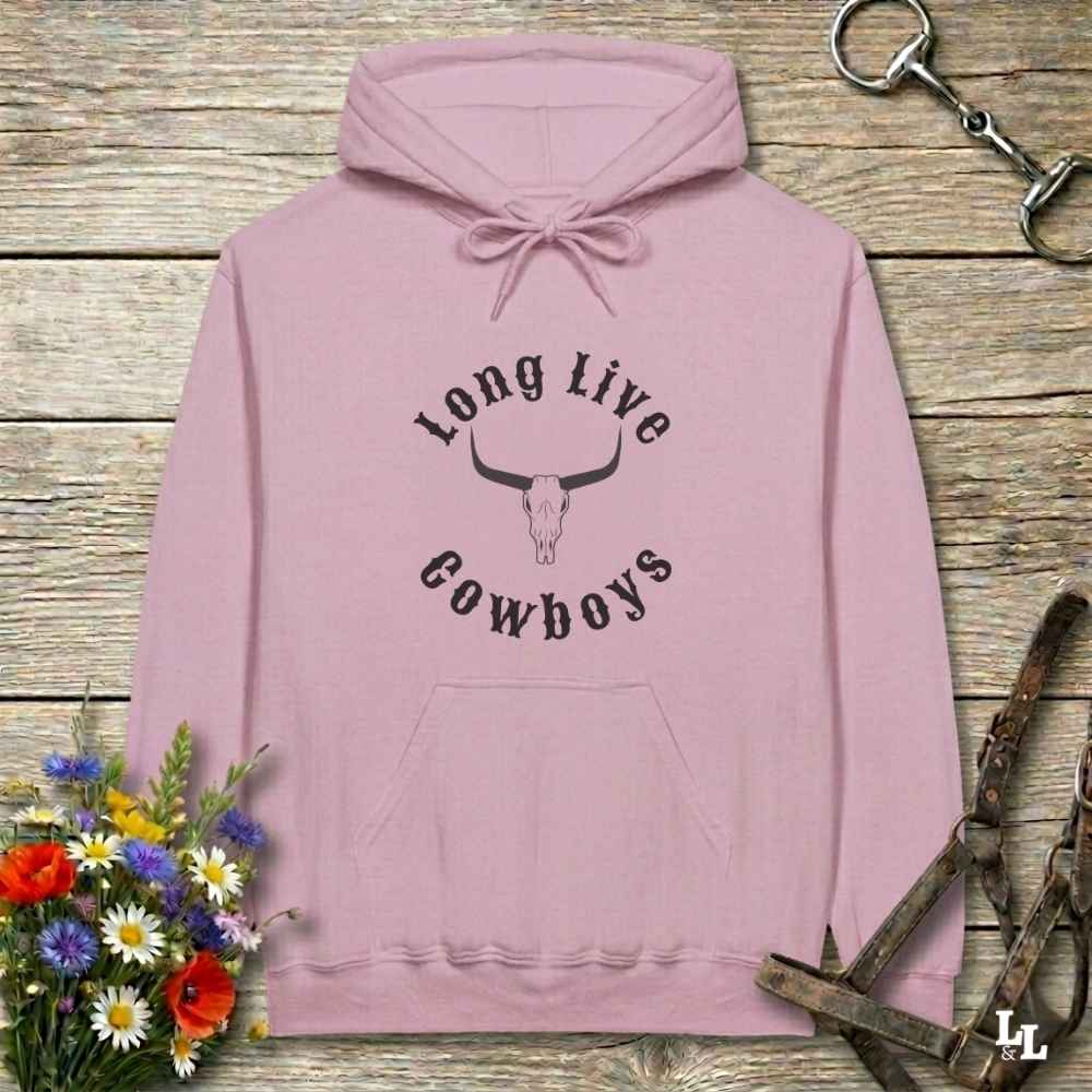 A dusty pink pullover hoodie with a black graphic print reading 'Long Live Cowboys' around a longhorn skull, laid flat on a rustic wooden surface with decorative flowers and horse tack props.