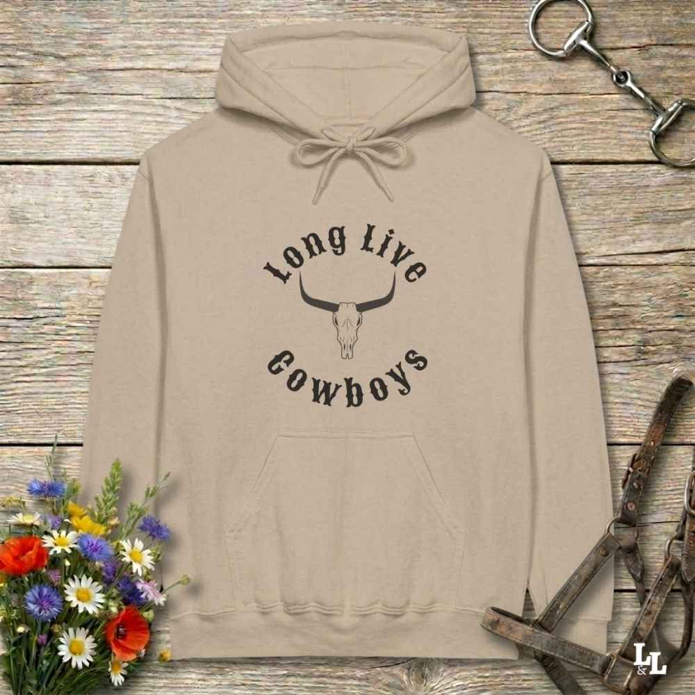 A beige pullover hoodie with a black graphic reading 'Long Live Cowboys' around a longhorn skull, laid flat on a rustic wood surface with flowers and horse tack props around it.