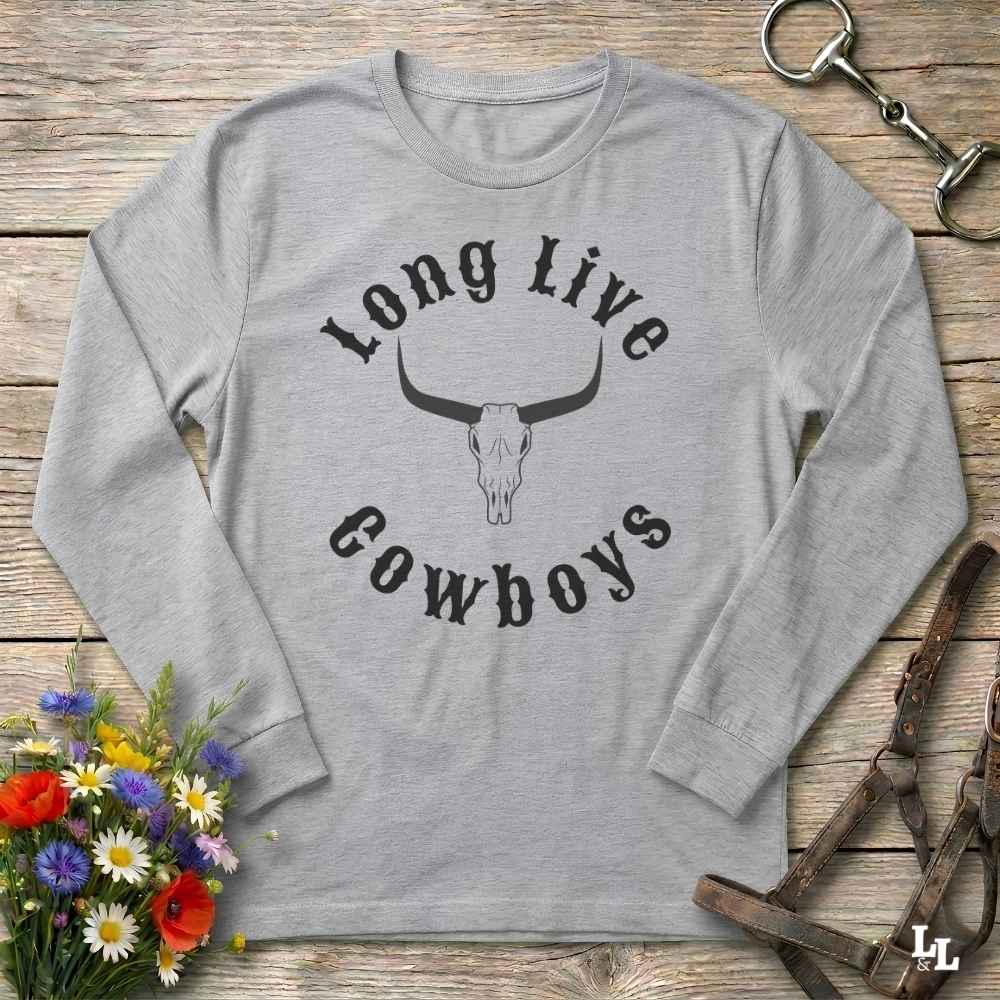 A light heather gray long-sleeve t-shirt with a black graphic print reading 'Long Live Cowboys' around a longhorn skull, laid flat on a rustic wooden surface with decorative flowers and leather tack around it.
