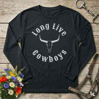 A black long-sleeve t-shirt with a white graphic reading 'Long Live Cowboys' around a longhorn skull, laid flat on a rustic wooden surface with flowers, a horse bit, and leather tack nearby.
