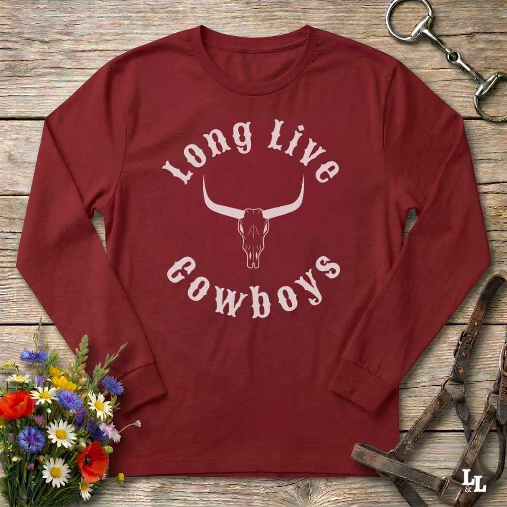 A long-sleeve crewneck shirt in dark red with a white graphic reading 'Long Live Cowboys' around a longhorn skull, laid flat on a rustic wood surface with flowers and horse tack props.
