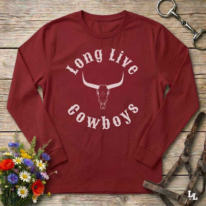 A long-sleeve crewneck shirt in dark red with a white graphic reading 'Long Live Cowboys' around a longhorn skull, laid flat on a rustic wood surface with flowers and horse tack props.