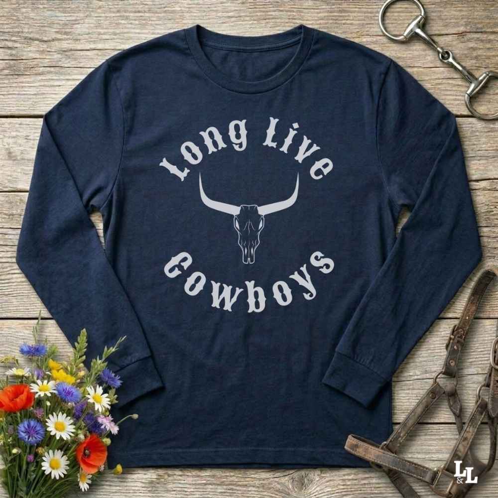 A navy blue long-sleeve t-shirt with a white graphic reading 'Long Live Cowboys' around a longhorn skull, laid flat on a rustic wooden surface with equestrian and floral props.