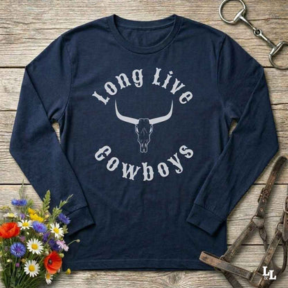 A navy blue long-sleeve t-shirt with a white graphic reading 'Long Live Cowboys' around a longhorn skull, laid flat on a rustic wooden surface with equestrian and floral props.