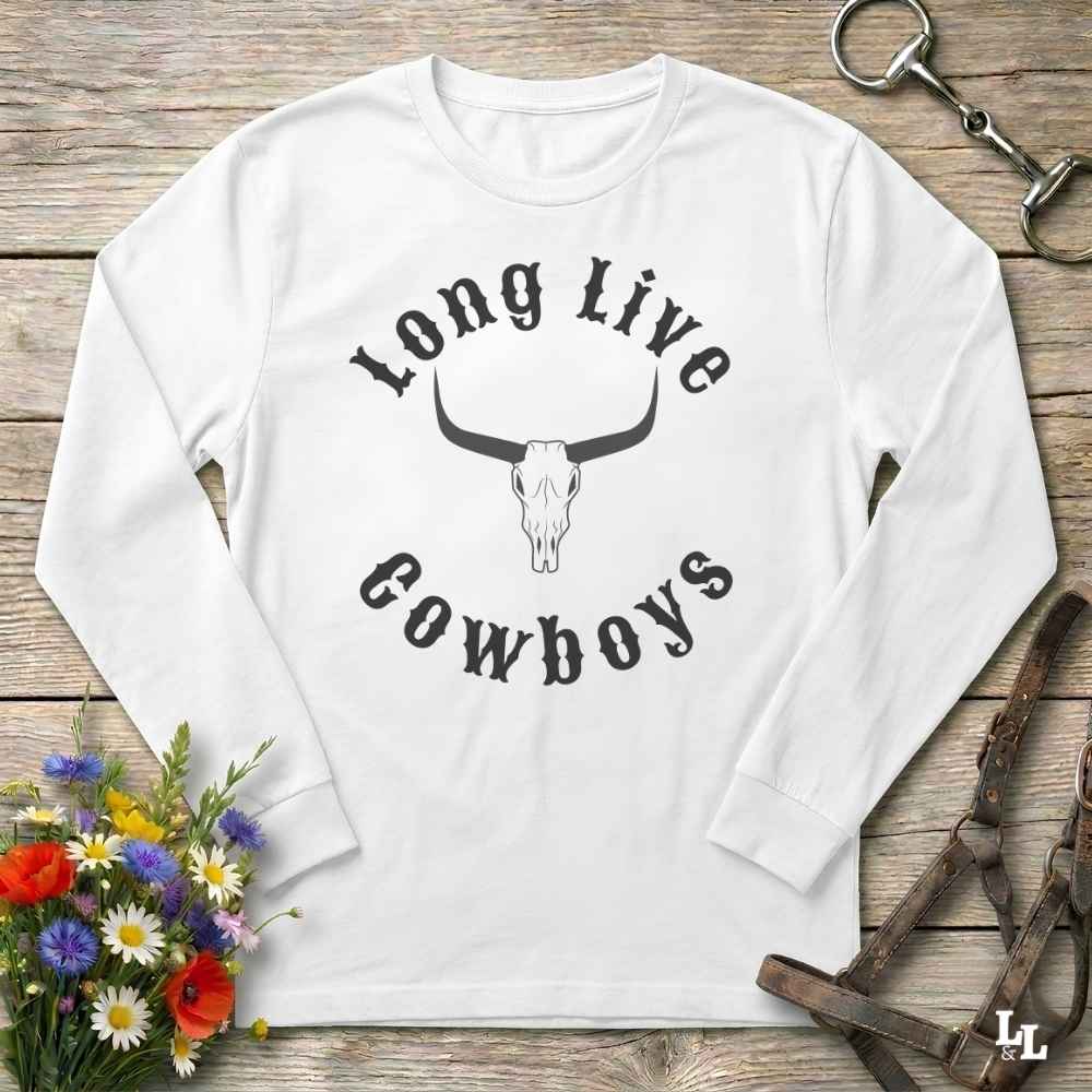 A white long-sleeve t-shirt with a black graphic reading 'Long Live Cowboys' around a longhorn skull, laid flat on a rustic wooden surface with flowers and horse tack props.