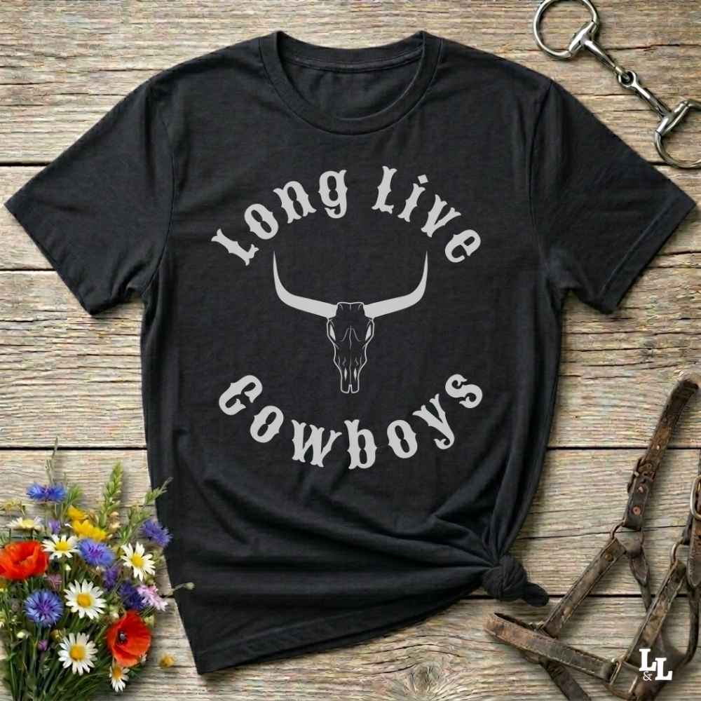 A black short-sleeve graphic T-shirt laid flat on a rustic wooden surface, with a white arched text design reading 'Long Live Cowboys' around a longhorn skull illustration. The shirt is styled with flowers, a horse bit, and leather tack around it.