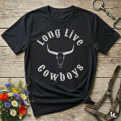 A black short-sleeve graphic T-shirt laid flat on a rustic wooden surface, with a white arched text design reading 'Long Live Cowboys' around a longhorn skull illustration. The shirt is styled with flowers, a horse bit, and leather tack around it.