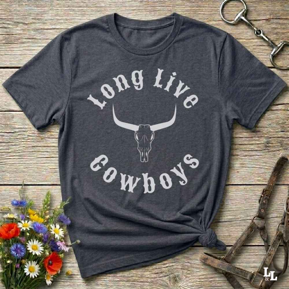A dark charcoal gray graphic t-shirt featuring a white longhorn skull and the words 'Long Live Cowboys' printed on the front. The shirt is styled in a flat-lay arrangement on a rustic wooden background, surrounded by decorative elements including a small bouquet of wildflowers, a metal keychain, and horse tack.