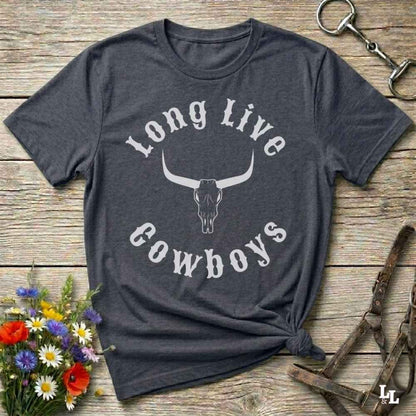 A dark charcoal gray graphic t-shirt featuring a white longhorn skull and the words 'Long Live Cowboys' printed on the front. The shirt is styled in a flat-lay arrangement on a rustic wooden background, surrounded by decorative elements including a small bouquet of wildflowers, a metal keychain, and horse tack.