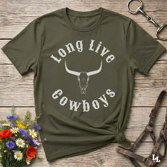 An olive green short-sleeve t-shirt with a white graphic reading 'Long Live Cowboys' around a longhorn skull, laid flat on a rustic wooden surface with flowers and leather tack props.