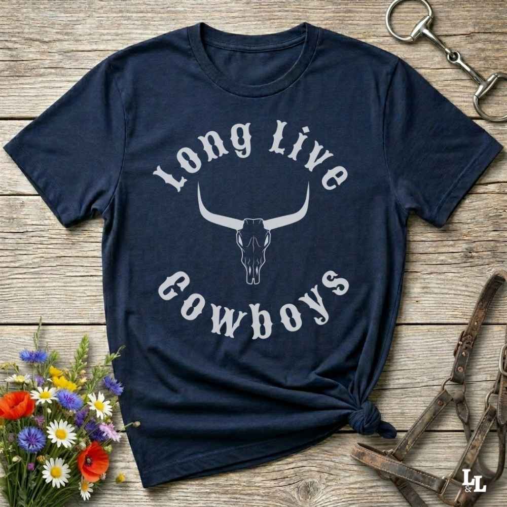 A navy blue short-sleeve t-shirt with a white graphic reading 'Long Live Cowboys' around a longhorn skull, laid flat on a rustic wooden surface with flowers and leather tack props.
