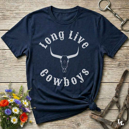 A navy blue short-sleeve t-shirt with a white graphic reading 'Long Live Cowboys' around a longhorn skull, laid flat on a rustic wooden surface with flowers and leather tack props.