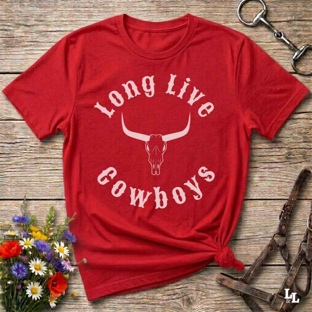 A red short-sleeve graphic T-shirt laid flat on a rustic wooden surface, with a white arched 'Long Live Cowboys' text design and a longhorn skull graphic on the front. The shirt is styled with flowers, a horse bit, and a leather bridle around it.