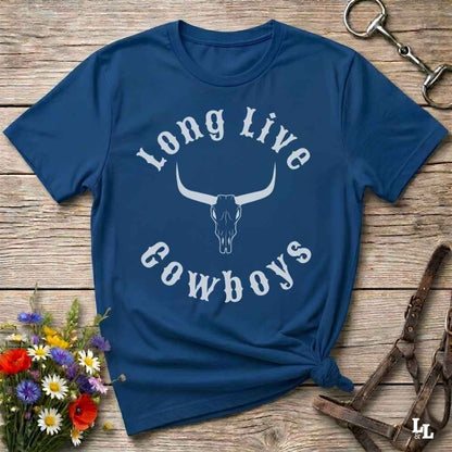 A navy blue short-sleeve t-shirt with a white graphic reading 'Long Live Cowboys' around a longhorn skull, laid flat on a rustic wood surface with flowers and horse tack props.