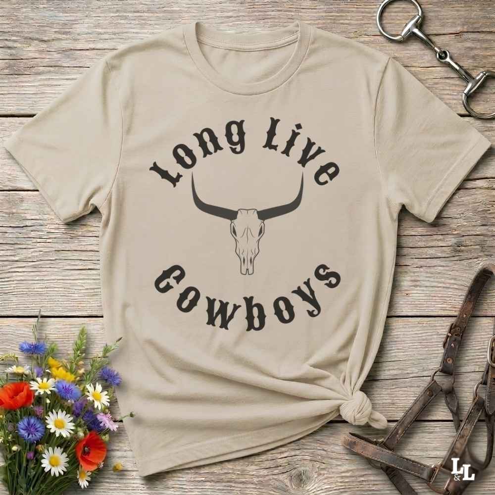 A beige graphic t-shirt with a black printed design reading 'Long Live Cowboys' around a bull skull, styled in a flat-lay on a rustic wooden background with flowers and leather tack.