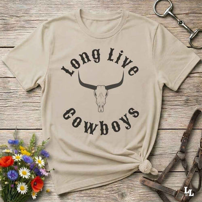 A beige graphic t-shirt with a black printed design reading 'Long Live Cowboys' around a bull skull, styled in a flat-lay on a rustic wooden background with flowers and leather tack.