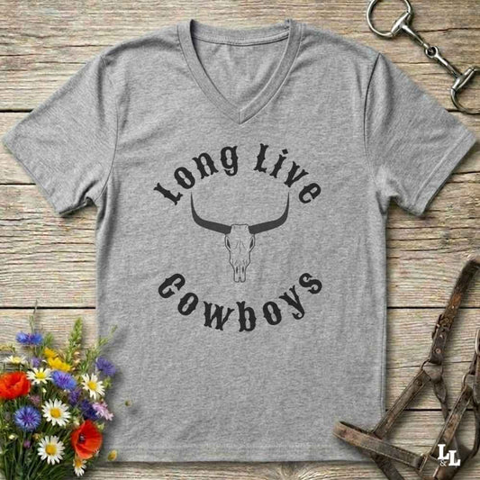 A heather gray V-neck t-shirt with a black graphic reading 'Long Live Cowboys' around a longhorn skull, laid flat on a rustic wood background with flowers and horse tack props.