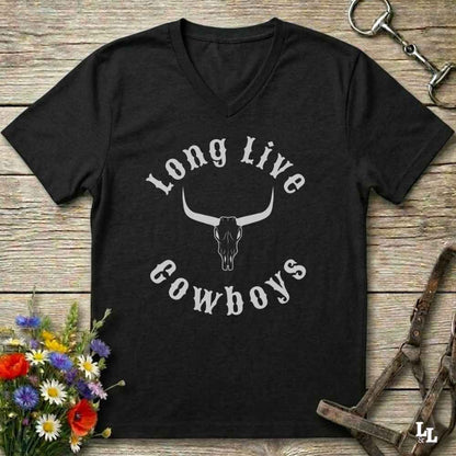 A black V-neck t-shirt with a white graphic reading 'Long Live Cowboys' around a longhorn skull, laid flat on a rustic wood background with flowers and horse tack props.