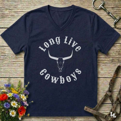 A navy blue V-neck t-shirt with a white graphic reading 'Long Live Cowboys' around a longhorn skull, laid flat on a rustic wooden surface with flowers and horse tack props.
