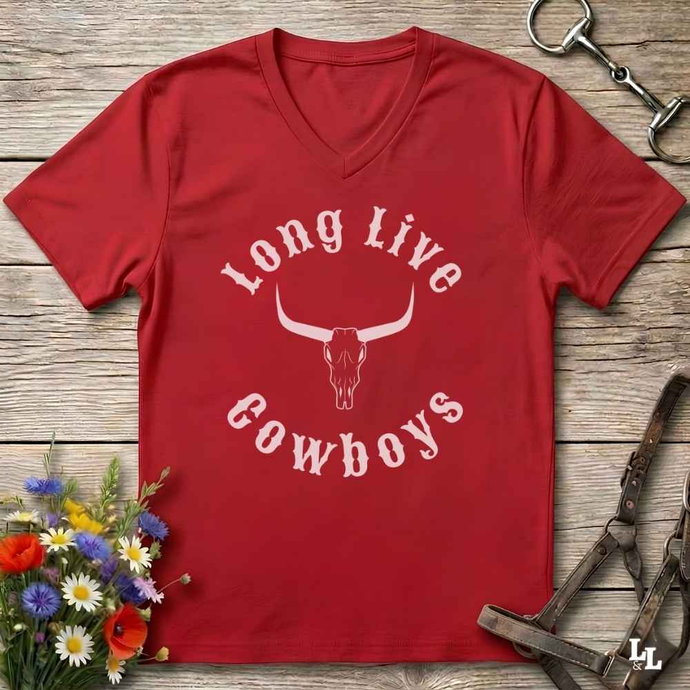 A red V-neck t-shirt with a white graphic reading 'Long Live Cowboys' around a longhorn skull, laid flat on a rustic wood background with flowers and horse tack props.
