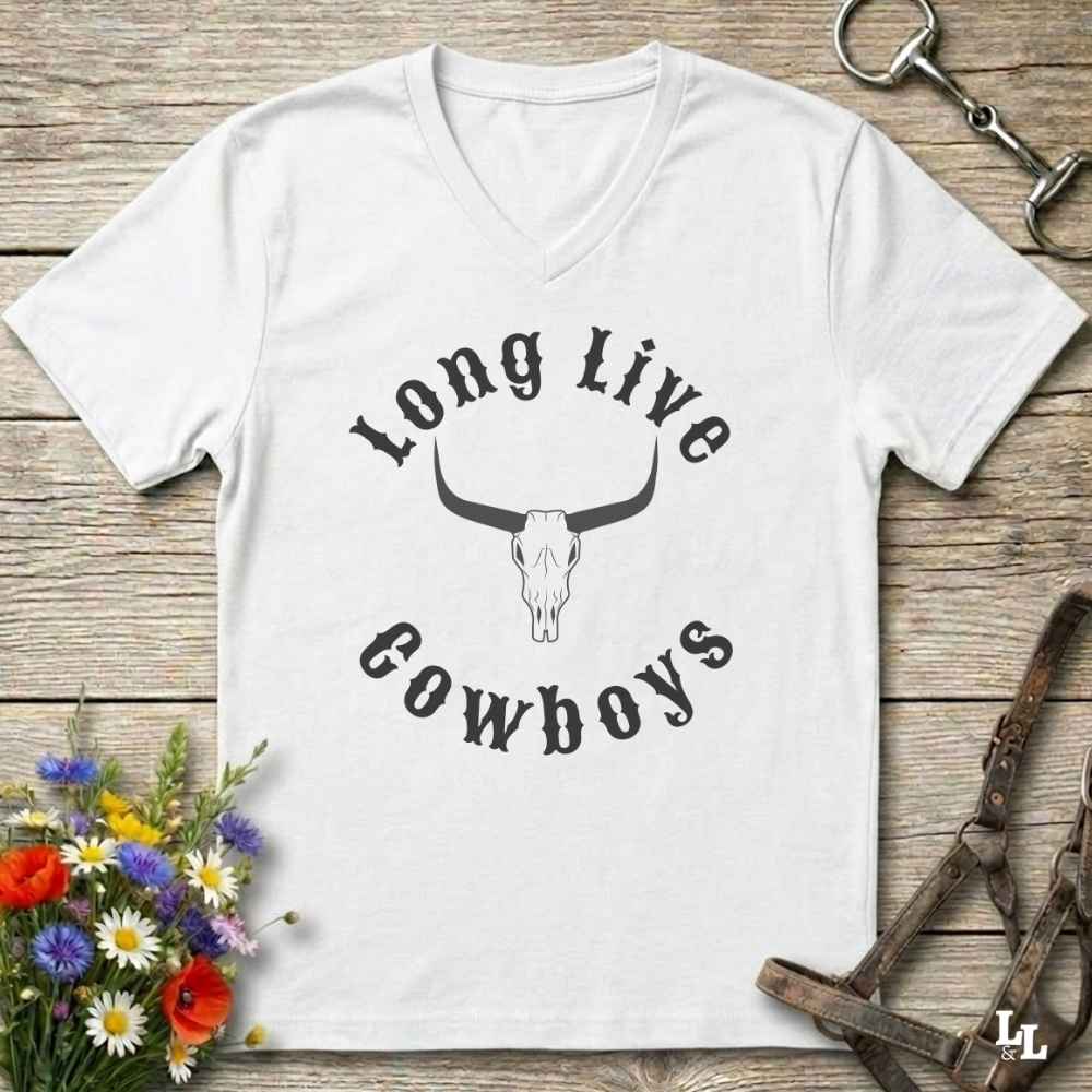 A white V-neck t-shirt with a black graphic reading 'Long Live Cowboys' around a longhorn skull, laid flat on a rustic wooden surface with flowers and horse tack props.