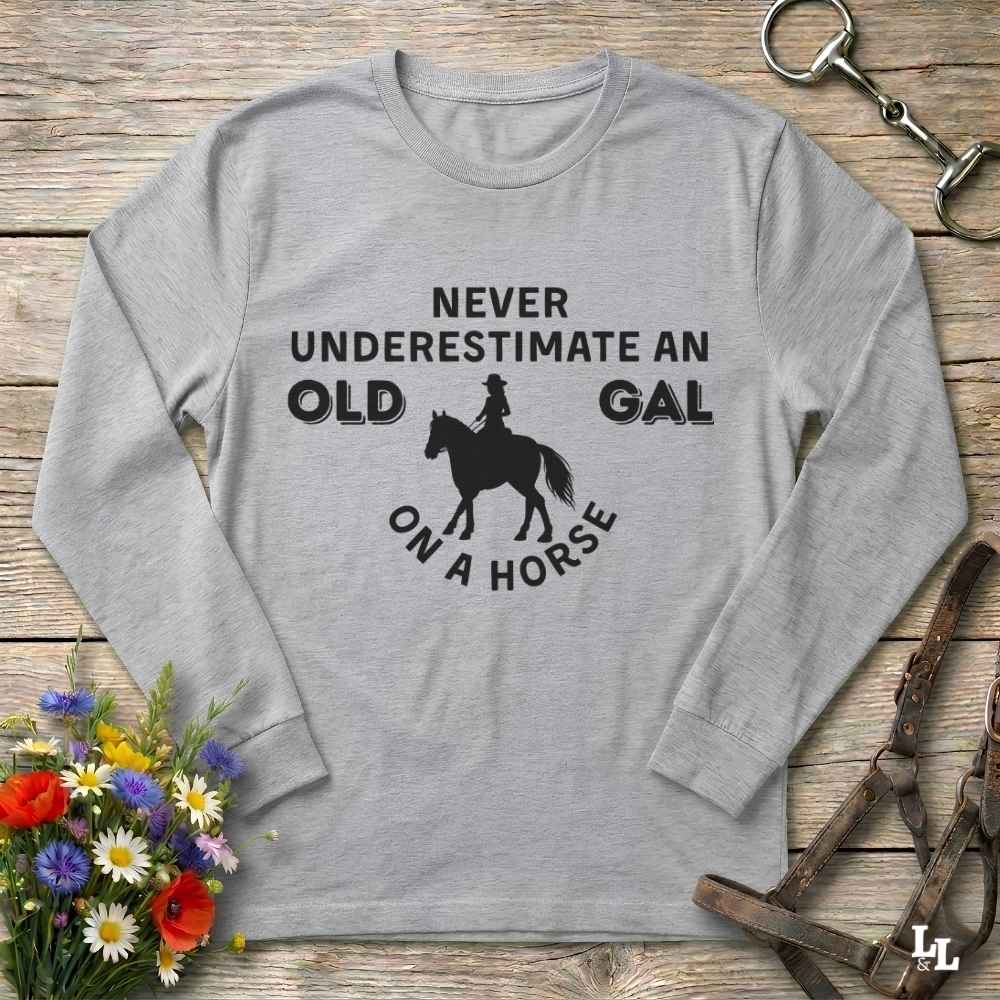A light gray long-sleeve t-shirt with a black graphic print that reads 'NEVER UNDERESTIMATE AN OLD GAL ON A HORSE' with a silhouette of a rider on a horse. The shirt is displayed flat on a rustic wooden background with equestrian-themed props including a leather bridle, horse bit, and a small bouquet of wildflowers.