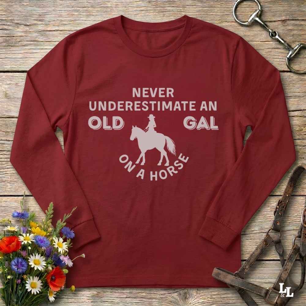 A long-sleeve crewneck t-shirt in maroon red with a white graphic print that reads 'NEVER UNDERESTIMATE AN OLD GAL ON A HORSE' surrounding a silhouette of a rider on a horse. The shirt is displayed flat on a rustic wooden background with equestrian-themed props including a horse bit, leather bridle, and a small bouquet of wildflowers.
