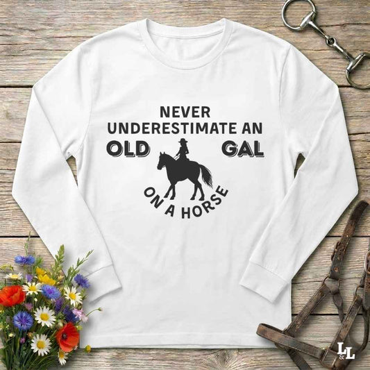 A white long-sleeve t-shirt with a black graphic print reading 'NEVER UNDERESTIMATE AN OLD GAL ON A HORSE' around a horse silhouette, displayed on a rustic wooden background with equestrian accessories and flowers.