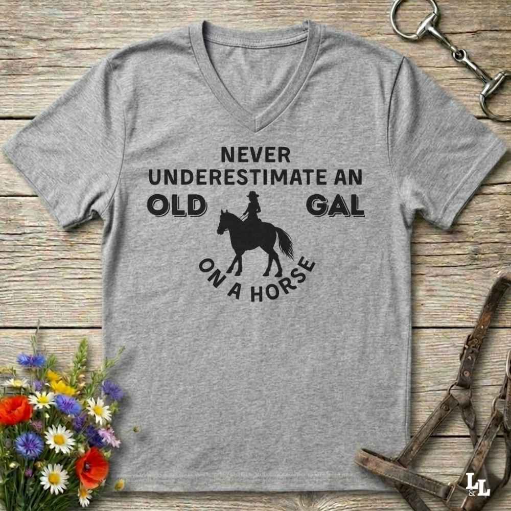 A heather gray V-neck t-shirt with a black graphic print that reads 'NEVER UNDERESTIMATE AN OLD GAL ON A HORSE' with a horse and rider silhouette, displayed flat on a rustic wooden background with equestrian accessories and flowers.