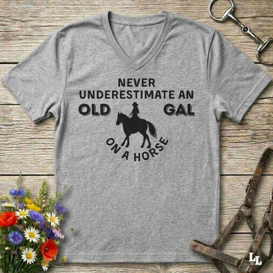 A heather gray V-neck t-shirt with a black graphic print that reads 'NEVER UNDERESTIMATE AN OLD GAL ON A HORSE' with a horse and rider silhouette, displayed flat on a rustic wooden background with equestrian accessories and flowers.