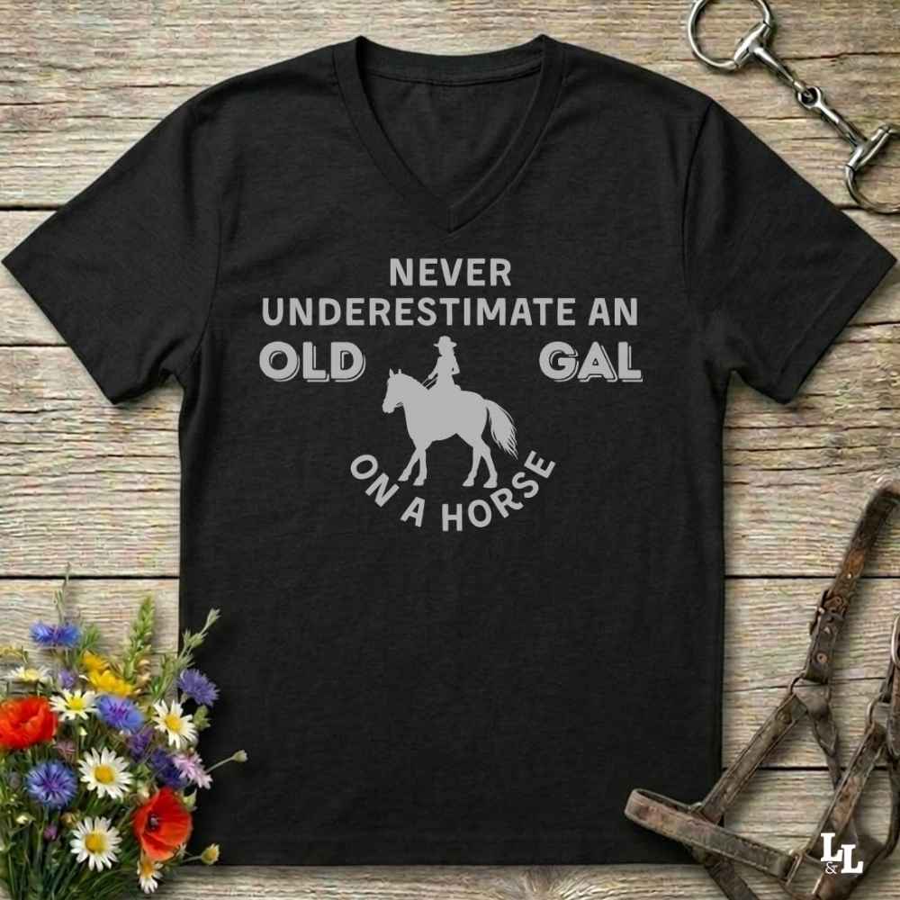 A black V-neck t-shirt with a white graphic print that reads 'NEVER UNDERESTIMATE AN OLD GAL ON A HORSE' surrounding a horse and rider silhouette. The shirt is displayed flat on a rustic wooden background with equestrian accessories and flowers.