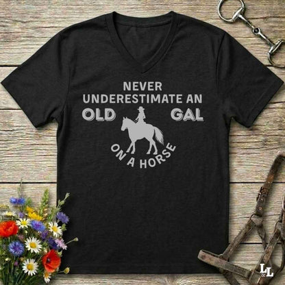 A black V-neck t-shirt with a white graphic print that reads 'NEVER UNDERESTIMATE AN OLD GAL ON A HORSE' surrounding a horse and rider silhouette. The shirt is displayed flat on a rustic wooden background with equestrian accessories and flowers.