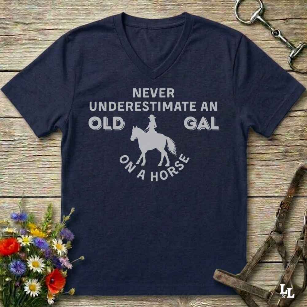 A navy blue V-neck t-shirt with a white graphic print that reads 'NEVER UNDERESTIMATE AN OLD GAL ON A HORSE' surrounding a silhouette of a horse and rider. The shirt is laid flat on a rustic wooden background with equestrian accessories and flowers.