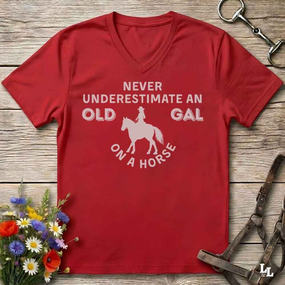 A red V-neck t-shirt with a white graphic print that reads 'NEVER UNDERESTIMATE AN OLD GAL ON A HORSE' with a silhouette of a rider on a horse. The shirt is displayed flat on a rustic wooden background with equestrian-themed props including a halter, tack, and a small bouquet of wildflowers.