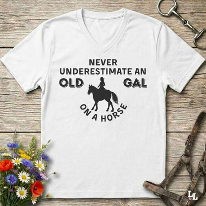A white V-neck t-shirt with a black graphic print reading 'NEVER UNDERESTIMATE AN OLD GAL ON A HORSE' with a silhouette of a rider on a horse, displayed flat on a rustic wooden background with equestrian accessories and flowers.