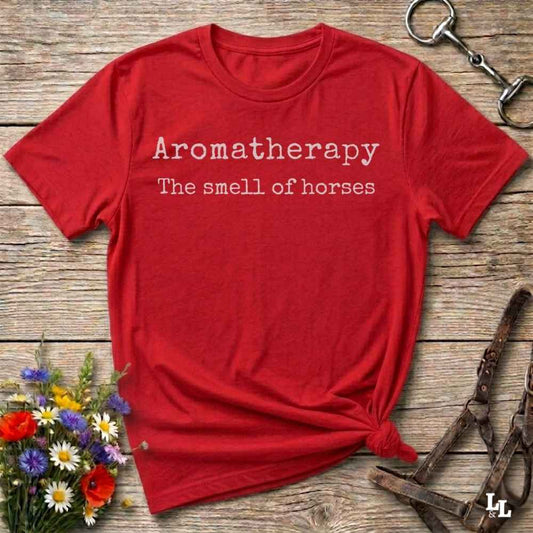 A red short-sleeve t-shirt with white text reading 'Aromatherapy The smell of horses' laid flat on a rustic wooden surface, styled with flowers, a horse bit, and a leather bridle.