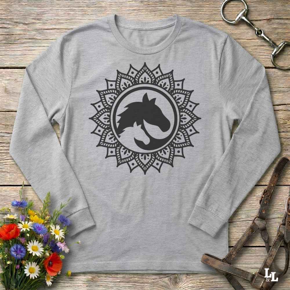 A long-sleeve crewneck shirt in grey, featuring a white graphic of a horse and cat silhouette inside a circular mandala design. The shirt is displayed flat on a rustic wooden background with equestrian tack and a small bouquet of flowers.