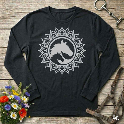 A long-sleeve crewneck shirt in black, featuring a white graphic of a horse and cat silhouette inside a circular mandala design. The shirt is displayed flat on a rustic wooden background with equestrian tack and a small bouquet of flowers.