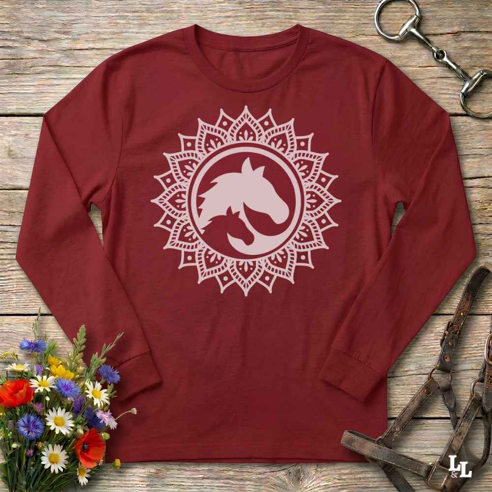 A long-sleeve crewneck shirt in maroon red, featuring a white graphic of a horse and cat silhouette inside a circular mandala design. The shirt is displayed flat on a rustic wooden background with equestrian tack and a small bouquet of flowers.