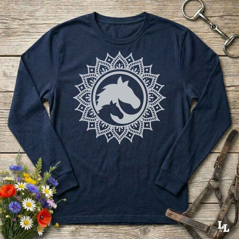 A long-sleeve crewneck shirt in navy blue , featuring a white graphic of a horse and cat silhouette inside a circular mandala design. The shirt is displayed flat on a rustic wooden background with equestrian tack and a small bouquet of flowers.